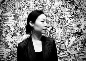A person stands calmly against a swirling, abstract black-and-white backdrop. The scene conveys a sense of contemplation and artistic intrigue.