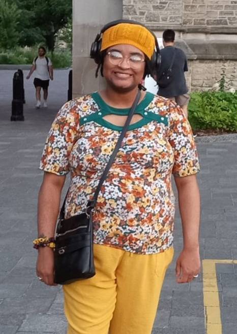 Person standing on a city street wearing a floral shirt, yellow pants, and sneakers, with a crossbody bag and a hat.