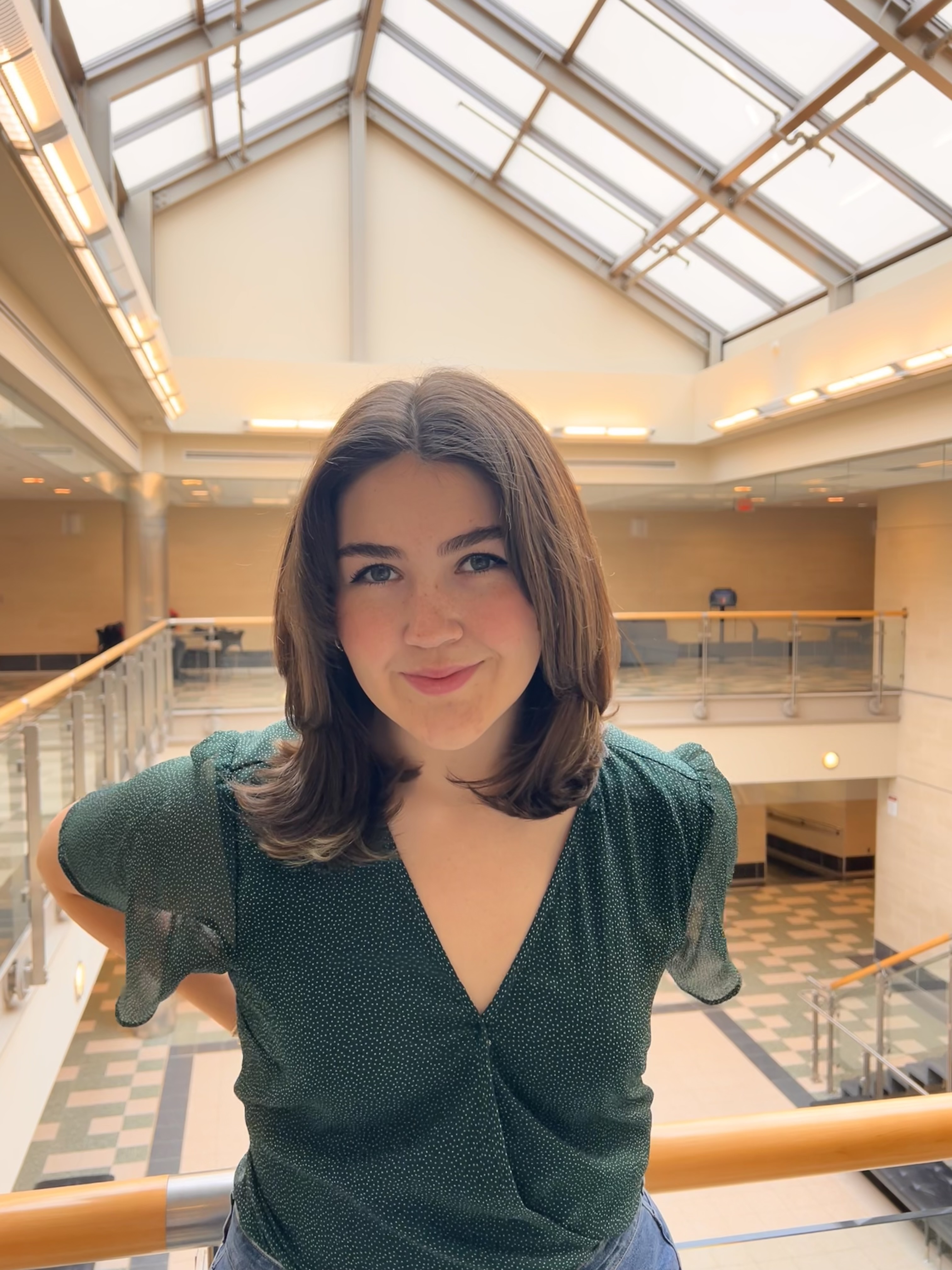 Person smiling for the camera inside a brightly lit atrium with a glass ceiling and railing.