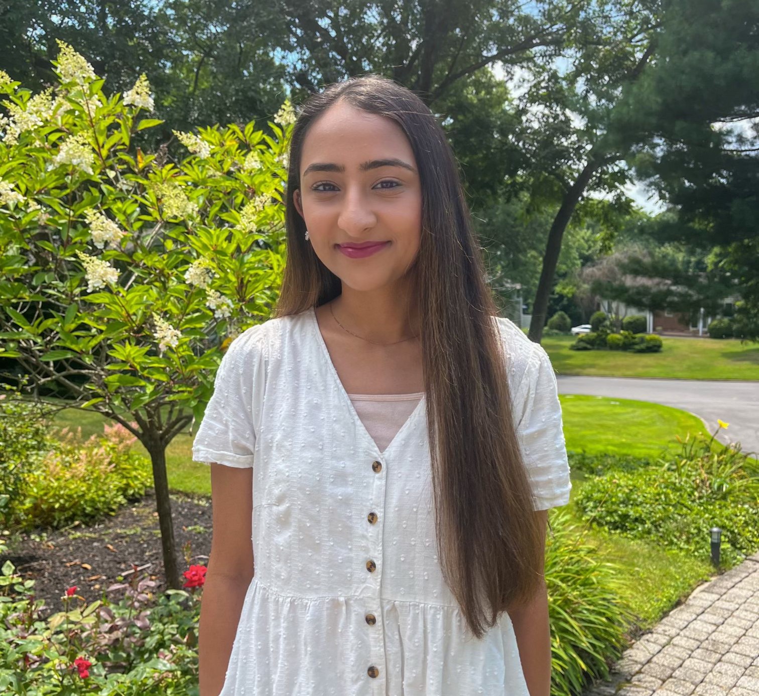 Person smiling in a white dress standing in a garden with green bushes and a pathway.