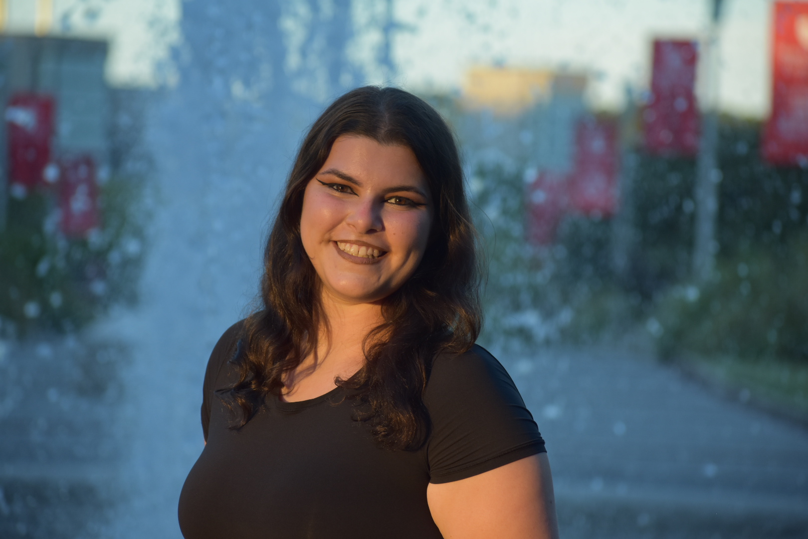 Smiling person standing in front of a gently splashing water fountain, bathed in sunlight.