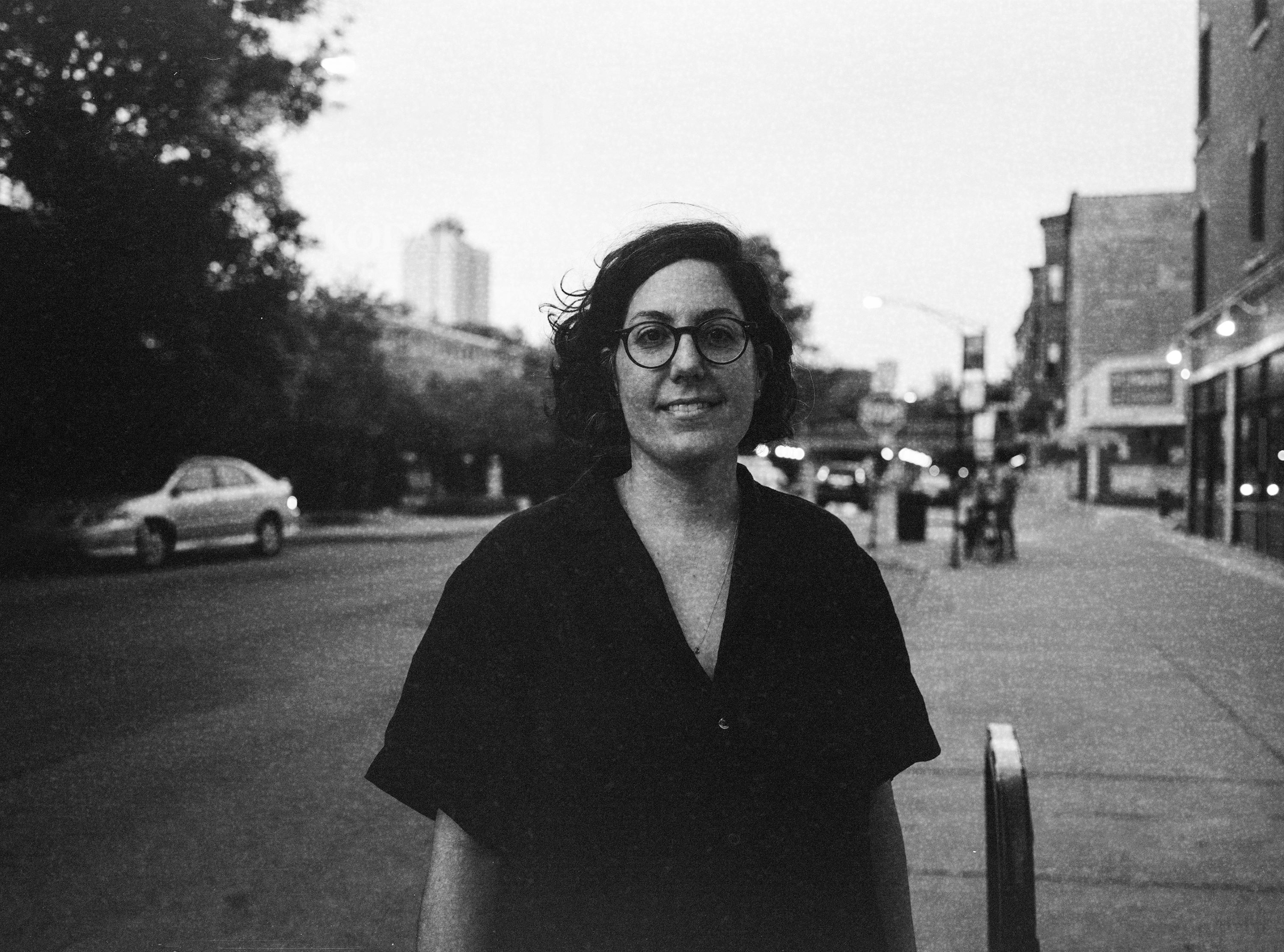 Smiling person wearing glasses, standing on an urban street with buildings and cars in the background.