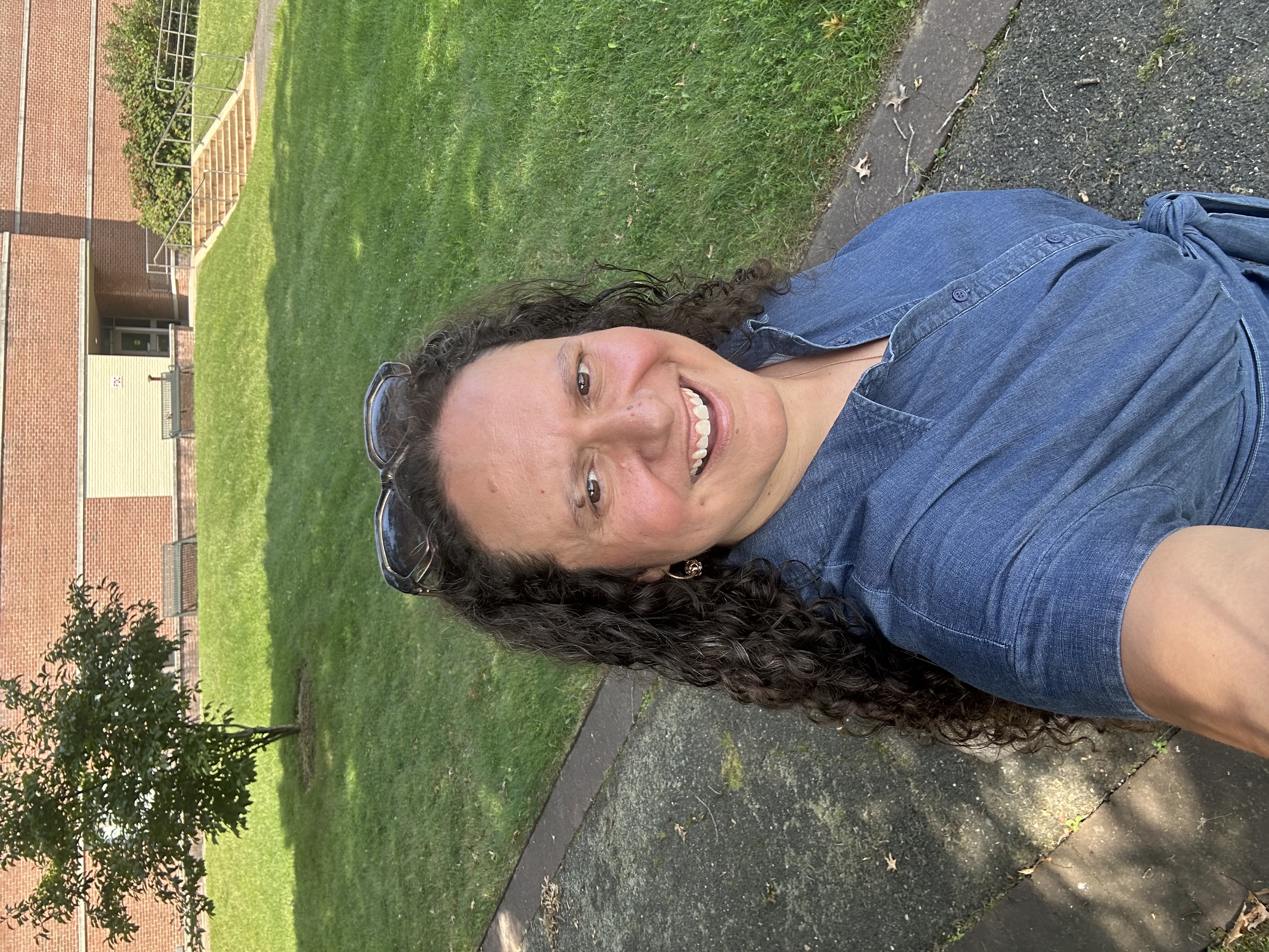 Person smiling outdoors with sunglasses on their head, and a building and trees in the background.