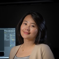 Portrait of a smiling person in a beige blazer, with a computer screen showing graphs in the background.