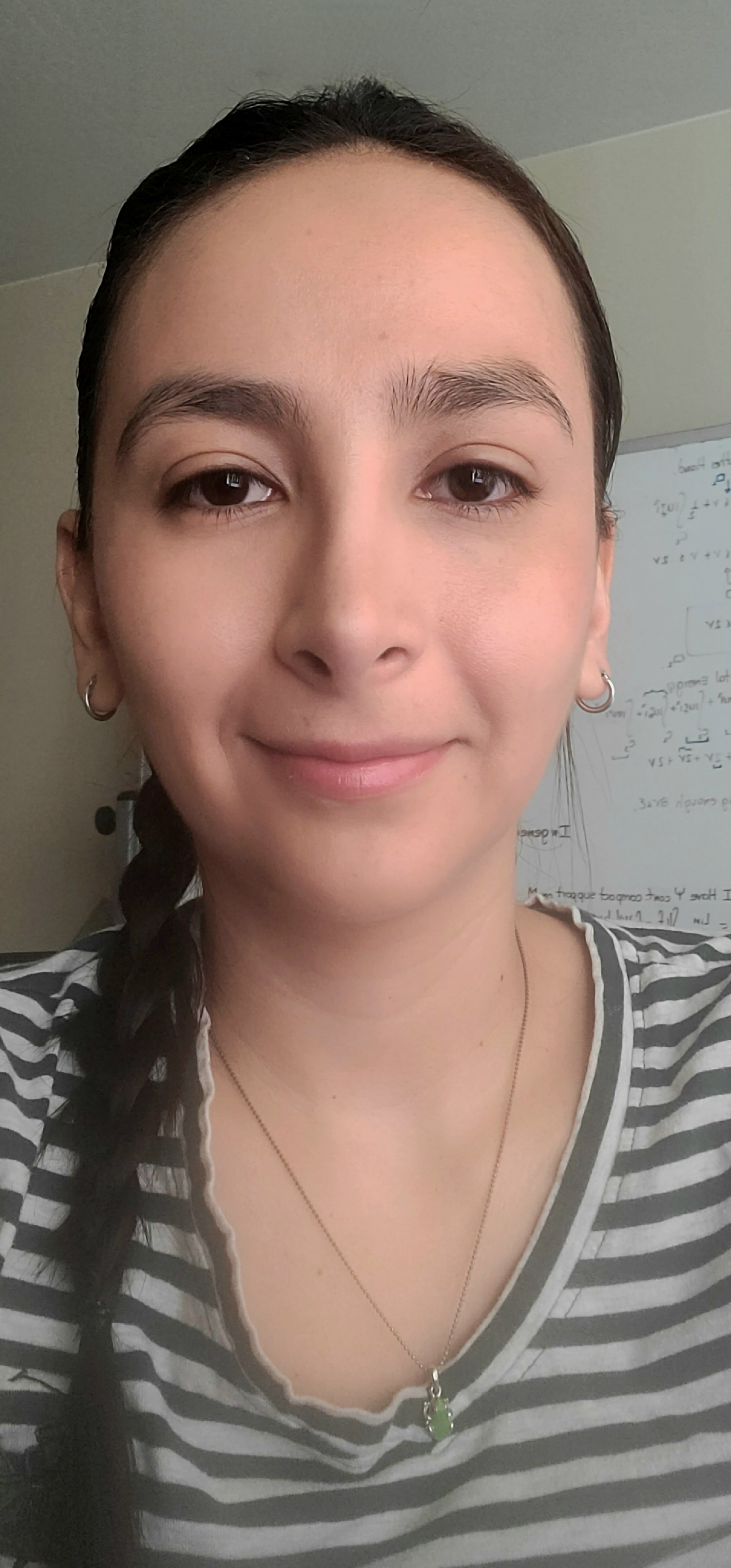 Person with a subtle smile, wearing a striped top and a jade pendant, standing in a room with a whiteboard covered in mathematical equations in the background.