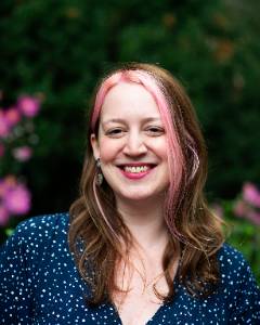 Person smiling in a blue polka dot top, with pink highlights in their hair, standing in front of green foliage with purple flowers.