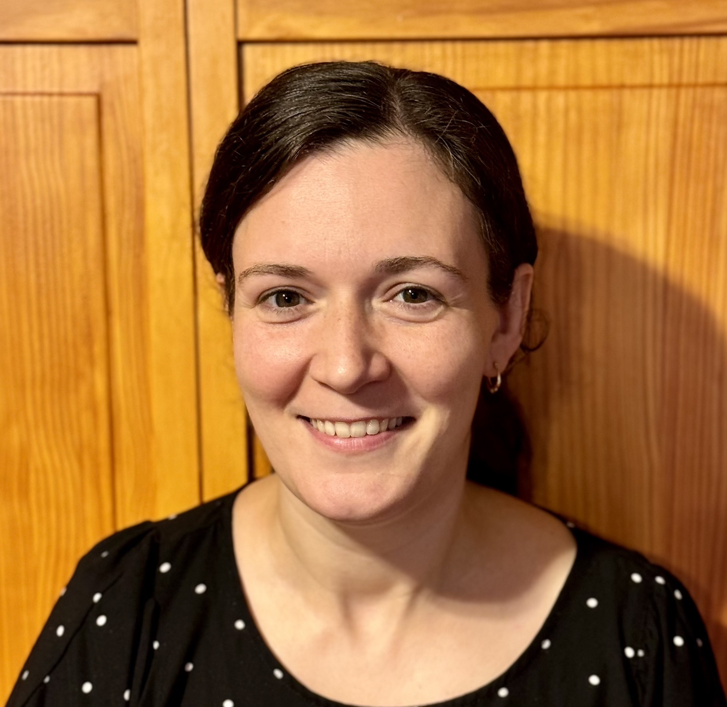 Close-up portrait of a smiling individual with short dark hair, wearing a polka dot top, against a wooden background.