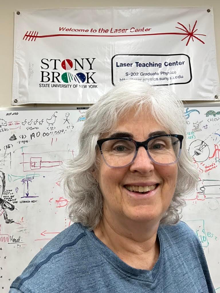 Individual smiling in front of a 'Welcome to the Laser Center' sign at Stony Brook University, with a whiteboard covered in colorful mathematical equations in the background.