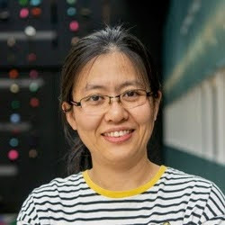 A portrait of an individual wearing glasses and a striped shirt, smiling in front of a blurred background with colorful lights.