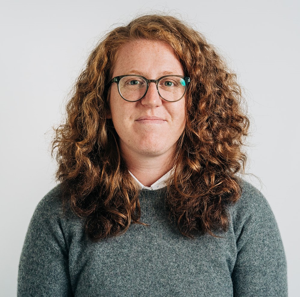 A woman with curly hair and glasses smiles warmly, showcasing her unique style and personality in a bright setting.