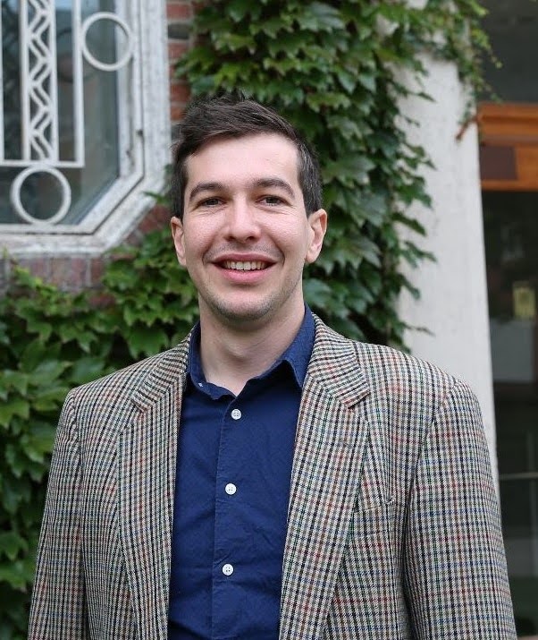A person smiling, standing outside a building covered with ivy. The person is wearing a plaid blazer over a blue shirt.