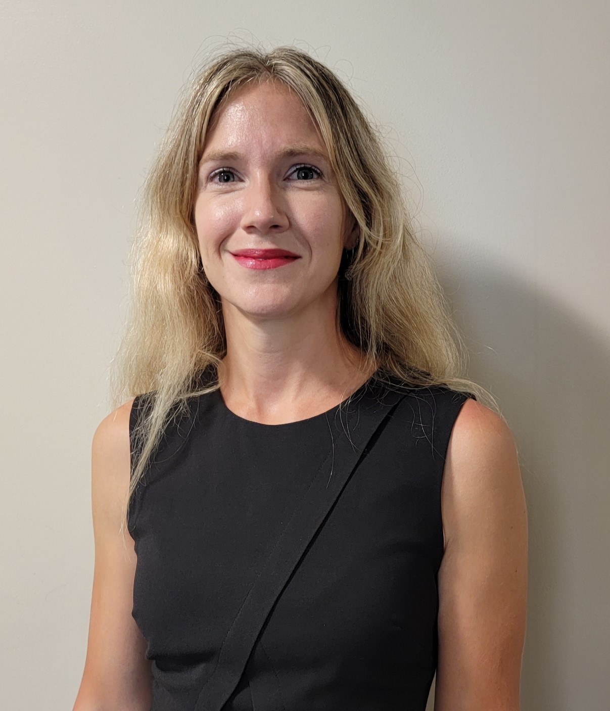 Portrait of an individual with shoulder-length blond hair, wearing a black sleeveless top, against a light-colored wall.