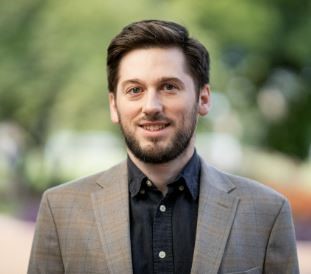 Portrait of a smiling person in a professional setting, wearing a gray blazer over a black shirt.