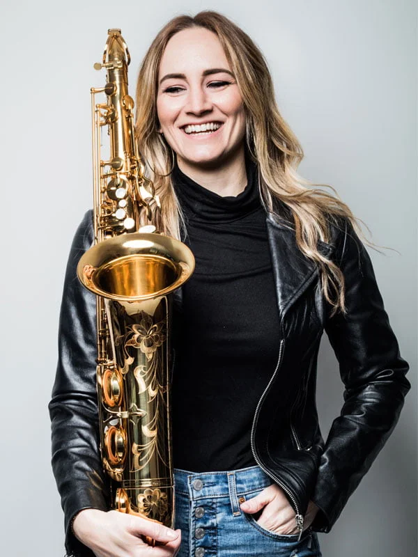 Person holding a saxophone, smiling, wearing a black turtleneck and leather jacket.