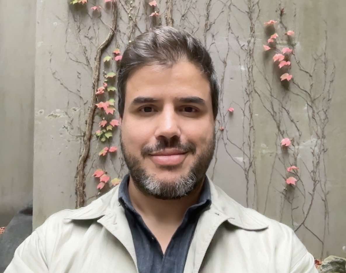 Person smiling at the camera in a tan jacket, standing in front of an architectural indoor background with flowers.