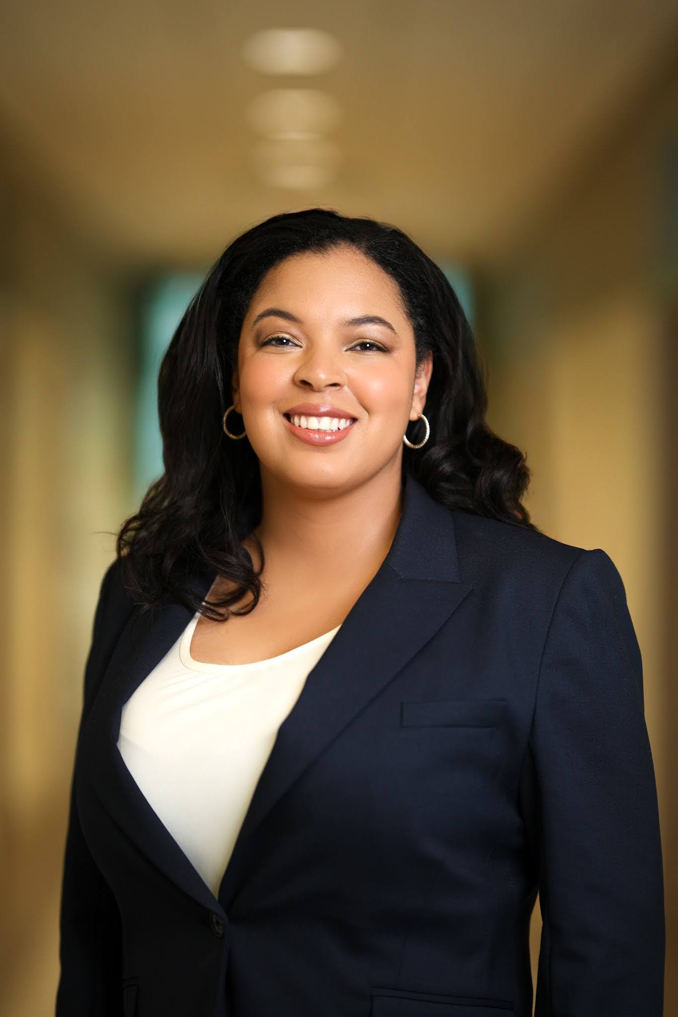 Professional portrait of a person smiling, wearing a dark suit and a light shirt, standing in a warmly lit corridor.