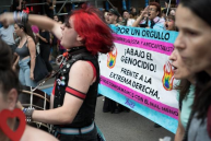 Participants holding a banner at an LGBTIQ+ and student pride march, with various signs and a crowd in the background.