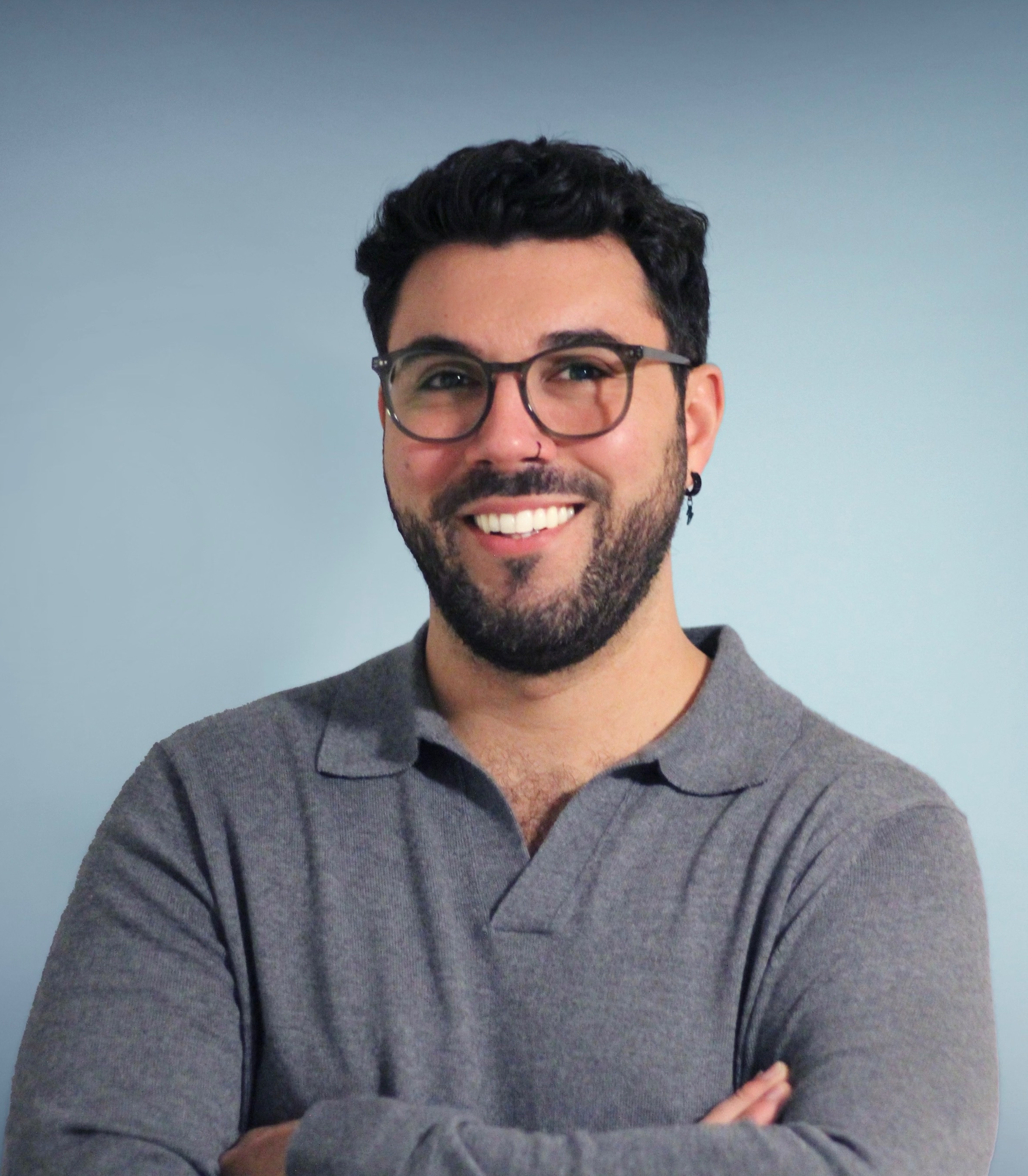 Stephan Brandt A smiling man with glasses and a beard stands against a light blue background. He is wearing a gray, long-sleeved, collared shirt and has his arms crossed."