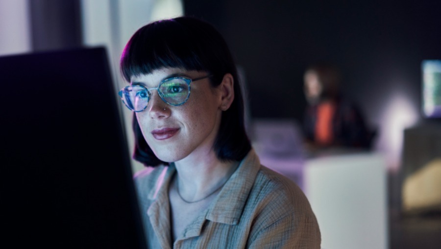 Woman sitting at a computer; digital code is reflected in her glasses.