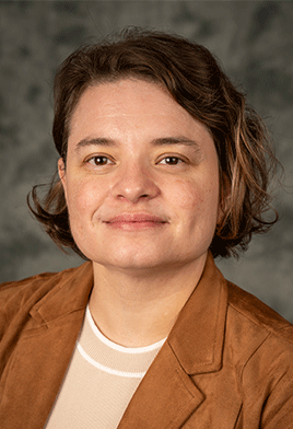 A person with short, wavy hair wearing a brown jacket and white top is smiling slightly against a gray, textured background, conveying a warm and approachable tone.