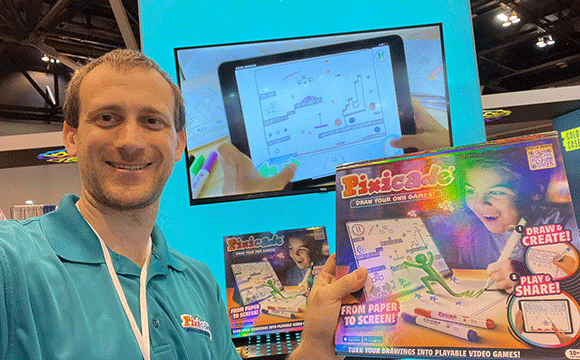 A man in a blue polo holds a Pixicade game box, smiling at a tech event. A screen in the background shows gameplay. The atmosphere is lively.