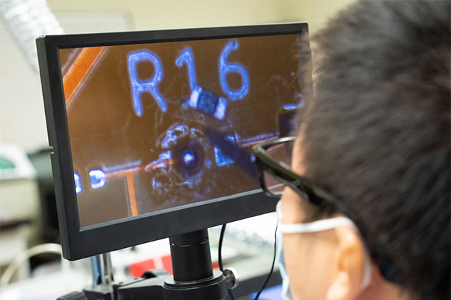 A person closely observes a monitor displaying magnified electronic components labeled "R16." The setting suggests careful examination or repair work.