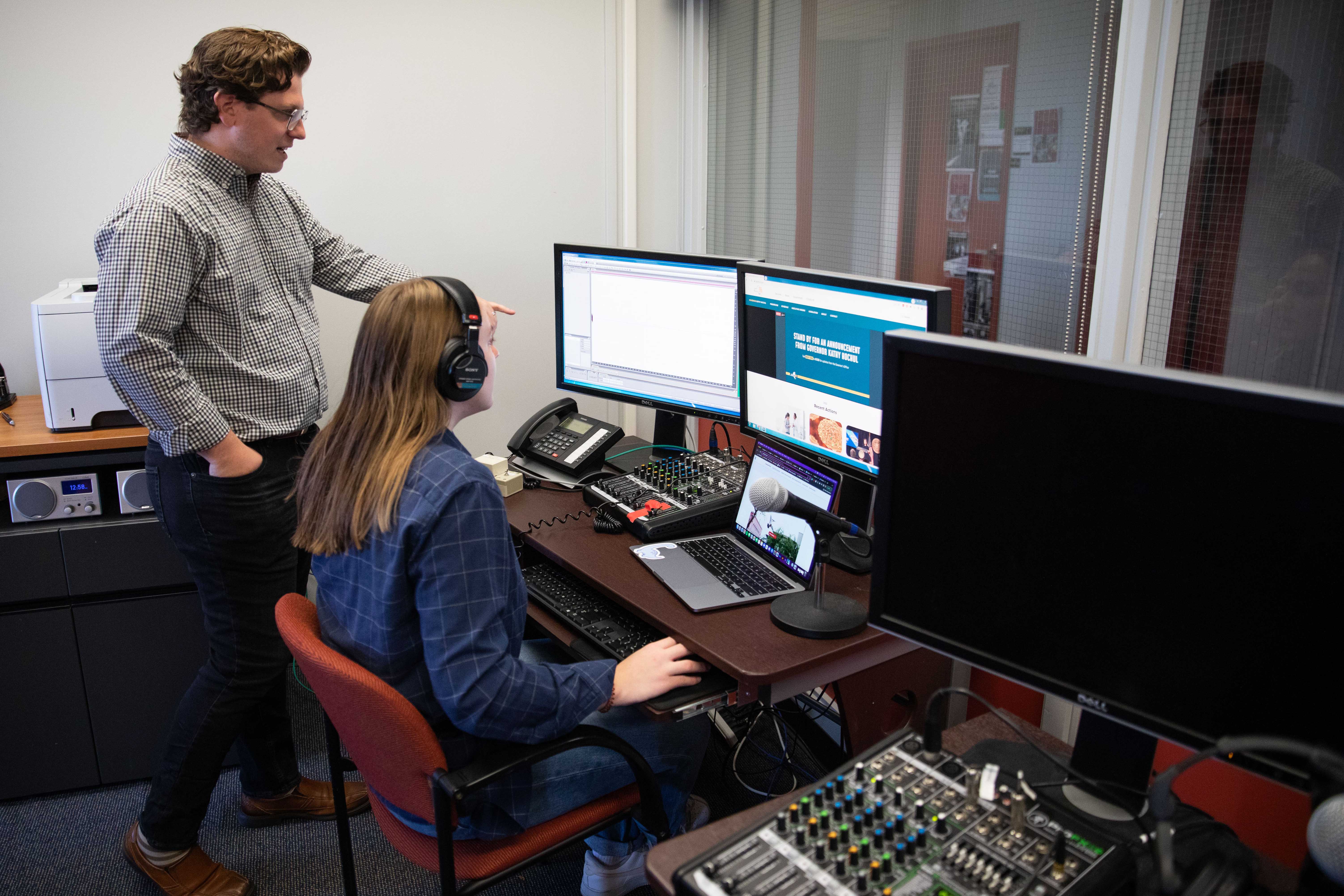 a student sits at a desk with an instructor standing next to her pointing at a computer screen