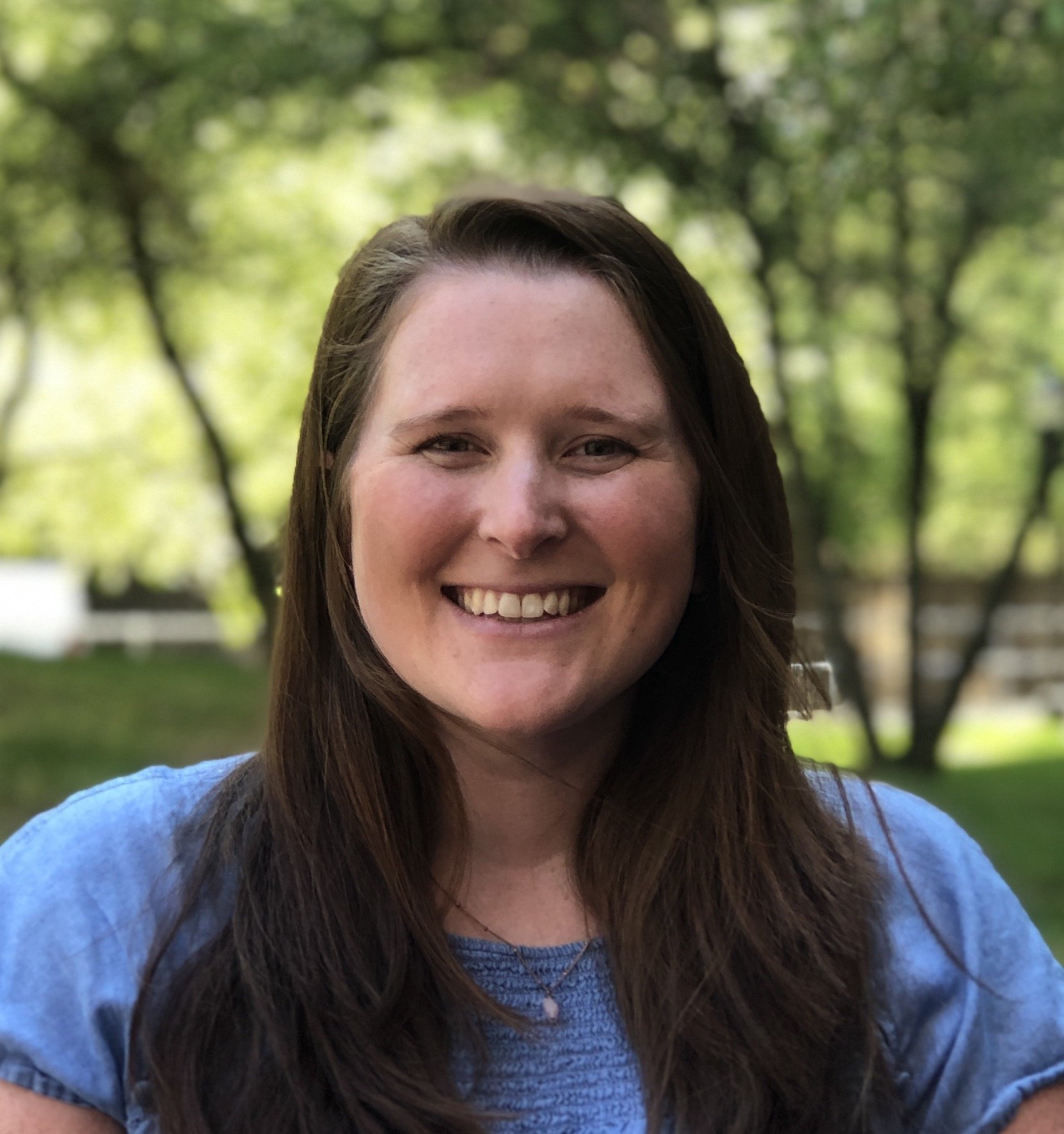 A smiling woman with long brown hair, wearing a blue textured top, stands in front of a soft-focus background of green trees and sunlight.
