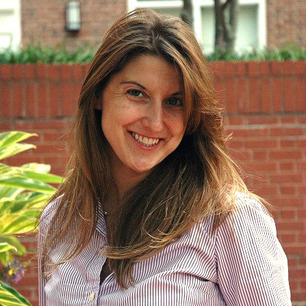 A smiling woman with long brown hair, wearing a pink and white striped button-down shirt, stands in front of a red brick wall. To her left, vibrant green and yellow leaves are visible.